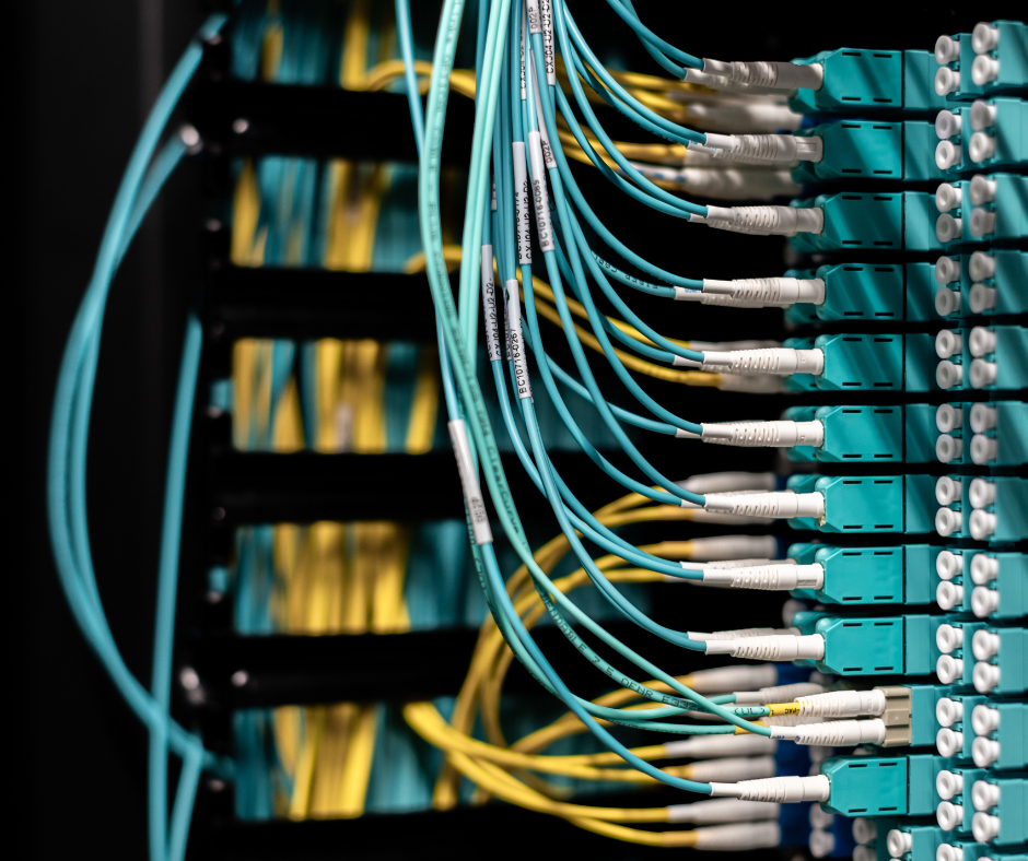A close-up view of networking equipment featuring numerous turquoise fiber optic cables plugged into rows of teal-colored modules on the right. In the background, yellow cables are visible, suggesting a complex cable management system within a server rack or similar setup. custom cable