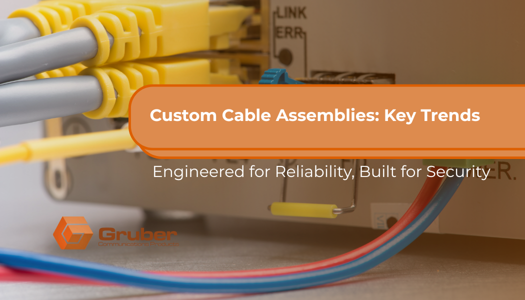 A close-up shot of several yellow ethernet cables plugged into a router or switch. In the foreground, out of focus, are red and blue cables. An orange overlay with rounded corners reads "Custom Cable Assemblies: Key Trends" in white text. Below it, in a smaller white font, is the phrase "Engineered for Reliability, Built for Security." In the bottom left corner, there is a logo that says "Gruber Communication Products" with an orange hexagonal graphic. custom cable