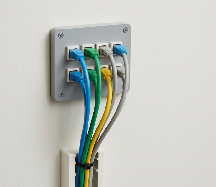 A square, gray, surface-mount box or Keystone wall plate secured to a white wall with four visible screws. It holds eight white keystone jacks, all of which are populated with colorful Ethernet patch cables. The cables (in blue, green, yellow, and gray) are neatly bundled with a cable tie as they exit the box through a cut-out below.